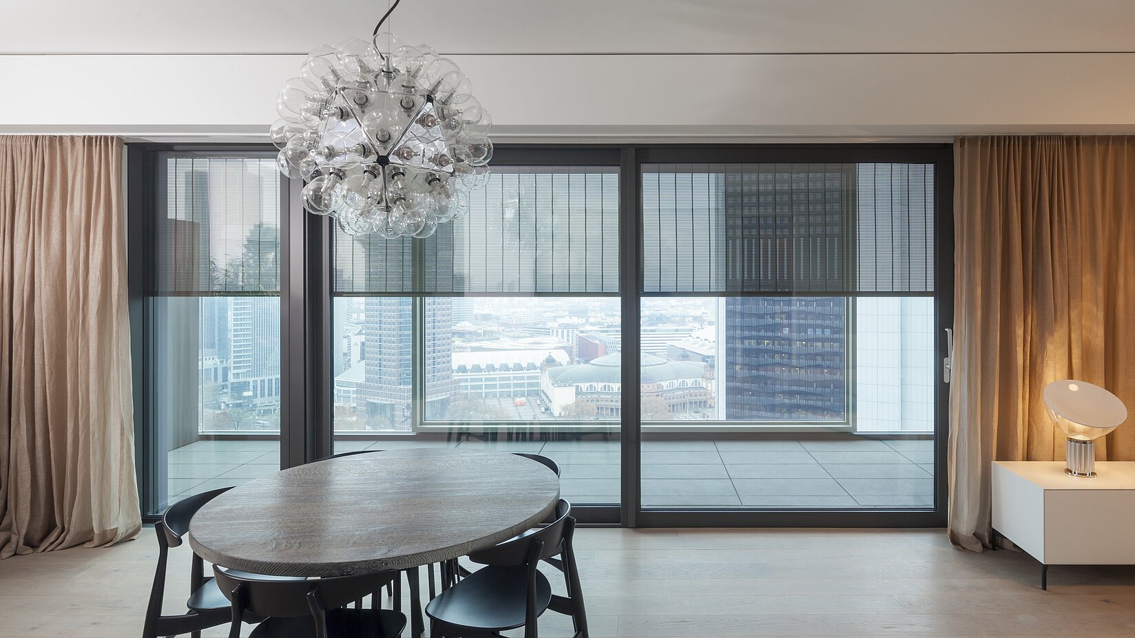 Essbereich mit grossem Holztisch vor Fensterfront mit MHZ S_enro Metallbehang und Ausblick auf Stadtgebäude.