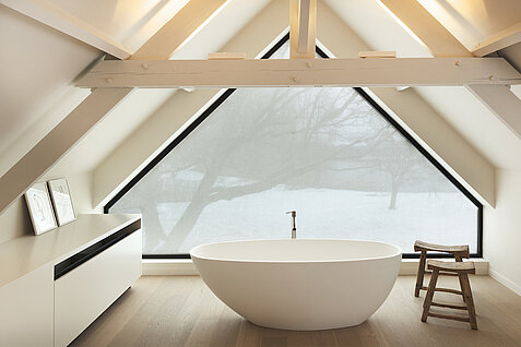 Badezimmer mit freistehender Badewanne vor einem grossen Dreiecksfenster mit Sichtschutztextil.