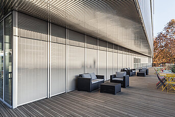 Terrasse mit modernen MHZ-Metallbehängen als vertikaler Sonnenschutz vor einer Glasfassade.