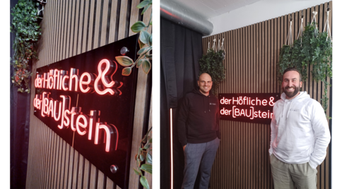 Zwei Männer stehen vor einer beleuchteten Neon-Schrift an einer Holzpaneelwand mit hängenden Pflanzen, der Text lautet „der Höfliche & der [BAU]stein“.