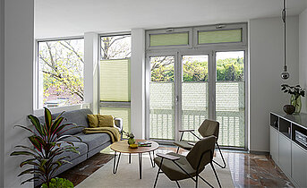 Wohnzimmer mit grünlich-transparenten MHZ DUETTE® Plissees vom Typ WILLOW an Fensterfront und Balkontür.