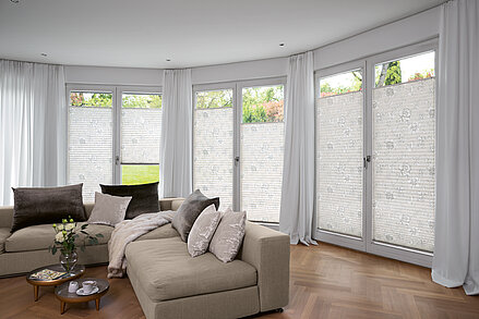 Florale Plissees in Weiss an bodentiefen Fenstern in elegantem Wohnzimmer mit Vorhängen.