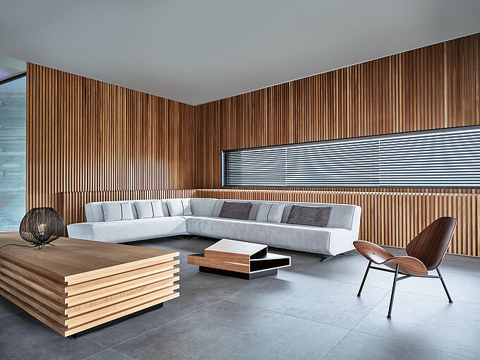 Modernes Wohnzimmer mit Holzlamellenwand, grauem Ecksofa und horizontaler Holzjalousie am schmalen Fenster.