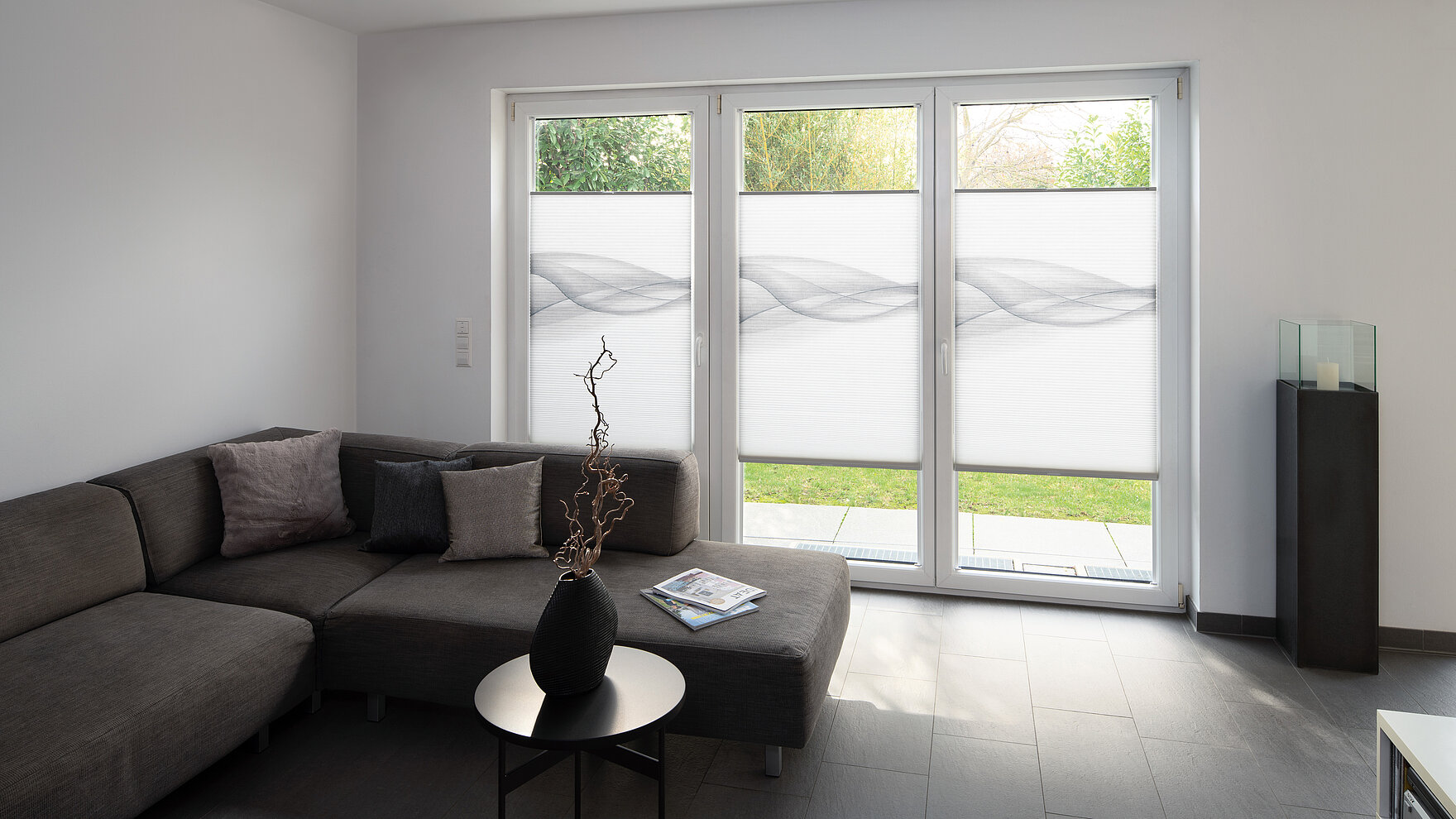 Wohnzimmer mit drei bodentiefen Fenstern, dekoriert mit grauen MHZ-Wabenplissees im Wellenmuster.